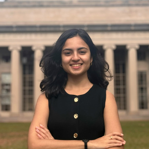 Priyanka Salunkhe with hands folded in front of her outside on MIT's Killian Court with building 10 and its large columns behind her