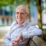 White man with white hair, smiling sitting on a park bench