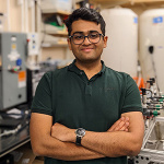 Aditya Ghodgaonkar standing in a lab facing camera with arms folded, smiling and wearing glasses
